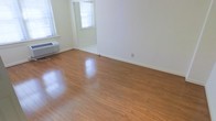 This single-floor space presents a clean, minimalist aesthetic defined by warm-toned hardwood flooring that runs throughout, lending a sense of continuity and warmth to the interior. The walls are finished in crisp white, creating a bright and airy atmosphere that feels open and welcoming. Natural light flows generously through multiple windows fitted with horizontal blinds, illuminating the polished floors and enhancing the overall sense of spaciousness. A wall-mounted PTAC unit provides in-unit climate control, offering year-round comfort. The layout is straightforward and versatile, with an open area that transitions through a subtle architectural threshold into an adjoining room or alcove, providing a degree of spatial definition while maintaining an open feel. The white baseboards and neutral color palette throughout offer a blank canvas well-suited to a variety of interior styles and furnishing arrangements. The space is practical and uncluttered, making it easy to envision and personalize for a range of uses. Overall, the combination of natural light, hardwood floors, and clean white finishes gives this space a bright, functional, and adaptable character.
