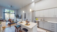 This well-designed two-bedroom, two-bathroom residence offers a thoughtfully organized single-story layout that maximizes its comfortable footprint. The open-concept kitchen and living area serves as the heart of the home, featuring soaring ceilings that amplify the sense of space. The kitchen is equipped with stainless steel appliances, white subway tile backsplash, two-tone grey and white cabinetry, and a central island with seating — ideal for casual dining or entertaining. Pendant lighting above the island adds a refined touch.

The living room flows naturally from the kitchen and opens to a private balcony through large sliding glass doors, framing lush tropical palm views and flooding the interior with natural light. Light-toned hardwood flooring runs throughout the main living areas, contributing to the bright, airy ambiance.

The primary bedroom features accent wall paneling, generous natural light from a large window, and ample closet storage. The second bedroom maintains a similarly clean, modern aesthetic. Each bathroom is finished with white quartz countertops and modern cabinetry; the primary bath includes a glass-enclosed shower, while the second bath offers a shower-tub combination.