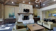 This single-story space showcases a compelling blend of rustic and contemporary design elements. The centerpiece of the room is a floor-to-ceiling stone fireplace, clad in natural limestone with a substantial reclaimed wood mantel and a sleek stainless steel firebox surround. A wall-mounted flat-screen television is integrated directly above the mantel, merging traditional craftsmanship with modern functionality.

The ceiling is a standout architectural feature, finished with reclaimed wood planking that adds warmth and texture while soaring to an impressive height. Recessed lighting and an industrial-style geometric chandelier illuminate the space, complementing the rustic overhead aesthetic.

The seating arrangement centers around a large, solid wood coffee table atop a woven area rug, flanked by plush olive-green sofas and armchairs that create an inviting, conversational layout. Stone accent walls add additional textural depth throughout the room.

Dark hardwood flooring anchors the space, providing a rich contrast to the lighter limestone tones. Large-format artwork in muted earth and teal tones adorns the walls, enhancing the overall warm and sophisticated ambiance of the space.