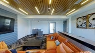 The Media Center is a thoughtfully designed single-floor space that blends comfort and contemporary style into a cohesive, functional environment. The most striking architectural feature is the slatted wood ceiling, which adds warmth and texture overhead while seamlessly integrating linear LED lighting strips that cast a clean, even glow throughout the space. The contrast between the natural wood tones of the ceiling and the light-colored walls creates a balanced and inviting atmosphere. The main lounge area is anchored by a plush, oversized sectional sofa in a bold burnt-orange upholstery, accented with decorative striped throw pillows that add a pop of pattern and color. A low-profile rectangular coffee table sits at the center of the seating arrangement, grounding the space and providing a practical surface. A patterned area rug beneath the furniture adds depth and visual interest to the flooring. Adjacent to the lounge area is a reception or service counter finished in a dark wood tone, complementing the ceiling's warm aesthetic. A secondary seating area with a sleek chair and side table is positioned near the perimeter, offering a more private spot for individual use. The space is finished with recessed lighting and HVAC vents that maintain a clean, modern ceiling line throughout.