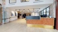 This single-floor space masterfully contrasts industrial heritage with modern design sensibilities. The entrance is defined by dramatically preserved original brick and weathered plaster walls, soaring double-height ceilings, and expansive black-framed windows that flood the space with natural light and offer street-level views of the surrounding urban environment. Red structural steel beams punctuate the rawness of the historic envelope, reinforcing the building's character.

At the heart of the lobby stands a thoughtfully designed reception desk featuring a warm terracotta herringbone wood-panelled base, a cool steel counter, and a polished brass toe kick — a refined focal point that balances old and new. Track lighting mounted on clean white ceilings provides flexible, gallery-quality illumination throughout.

Beyond the reception area, a lounge is furnished with modular pink upholstered seating arranged around natural wood coffee tables atop a bold navy patterned area rug. Wall-mounted shelving displays framed photographs and potted greenery, adding a personal, curated touch. A large graphic mural and exposed original painted surfaces contribute to a creative, heritage-rich ambiance. Light tile flooring ties all zones together cohesively throughout the space.