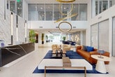 Located at 120 N Compass Way in Dania Beach, Florida, this striking amenity space spans a generous single floor and delivers an upscale, resort-inspired atmosphere throughout. The grand lobby entrance commands immediate attention with its soaring double-height ceilings and dramatic floor-to-ceiling glass walls that flood the interior with natural light and offer open views of the surrounding outdoor greenery and pool area. A sculptural gold ring chandelier serves as a defining centerpiece, hovering above a refined seating arrangement of curved velvet chairs, a rich cognac leather sofa, and deep navy area rugs atop large-format white porcelain tile flooring. A sleek reception desk anchors one side of the lobby, while lush tropical plants add organic warmth throughout. The space transitions seamlessly into a expansive social lounge, where oversized sectional sofas, a textured stone accent wall, and vibrant patterned throw pillows create an inviting, relaxed environment with direct sightlines to the outdoors. A fully equipped kitchen area features bold saffron-yellow walls, slate-grey cabinetry, a marble-slab backsplash, and a wood-trimmed island with a stainless steel sink. A formal dining area with a large communal table and sculptural pendant lighting completes the space, making it well-suited for both casual gatherings and private events.