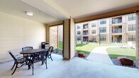 This covered outdoor amenity space is designed to serve as a central gathering hub within a modern residential community. The area is organized into several distinct zones, each offering a different seating experience. Ornate cast-iron round tables paired with matching chairs provide casual dining spots, while high-top bar tables with bar stools offer a more relaxed social setting. A dedicated lounge zone features dark contemporary sofas arranged around a coffee table atop a patterned area rug, creating a comfortable retreat for conversation or relaxation.

The architectural design features horizontal shiplap-style paneling in neutral tones, accented by warm wood-framed columns and stone-clad pillars that add texture and visual interest throughout the space. Flush-mount ceiling lights provide consistent illumination beneath the covered structure. The open sides of the space connect seamlessly to the surrounding grounds, allowing natural light and fresh air to flow freely through.

Views from the space look out onto a well-maintained green courtyard with landscaped planting beds on one side, and a pool area with a perimeter fence on the other. The multi-story buildings of the community frame the backdrop, reinforcing the sense of a cohesive, well-appointed residential environment. The overall ambiance is welcoming, versatile, and ideal for both quiet relaxation and social gatherings.