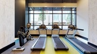 This contemporary fitness center is thoughtfully designed to support a full range of workout needs within a single, open-plan floor. The cardio zone is anchored by a row of treadmills and stationary bikes positioned directly in front of expansive floor-to-ceiling windows, flooding the space with natural light and offering views of the surrounding streetscape. Adjacent to this area, a collection of elliptical trainers and a rowing machine are arranged in front of a bold, colorful mosaic accent wall—a striking visual feature that energizes the space. Full-length mirrors amplify the sense of openness throughout. A dedicated mat and stretching zone features a blue rubberized floor, providing a safe, cushioned surface for functional training and recovery. A lounge nook with a built-in bench and decorative pillows offers a comfortable area to rest before or after a workout, bathed in natural light from large windows. The entry area includes a bank of dark metal lockers topped by a wall-mounted television for added convenience. The design palette blends dark ceilings, light stone-finish columns, and vibrant multi-toned carpeting, creating an atmosphere that is both dynamic and functional.