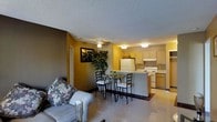 This single-floor space offers a practical and cohesive open-concept layout that seamlessly integrates the living, dining, and kitchen areas into one unified living environment. The interior is characterized by warm yellow walls that create an inviting and cheerful atmosphere throughout.

The living area is comfortably appointed and flows naturally into a compact dining space, which features a sleek glass-top table with black metal chairs — an elegant centerpiece for the room. A ceiling fan overhead provides both function and a subtle decorative touch, while a textured popcorn ceiling adds a classic finish to the space.

The kitchen is efficiently designed with light wood cabinetry, a white refrigerator, and a white electric range. A peninsula counter serves as both a workspace and a natural divider between the kitchen and the adjoining living and dining areas. A tile floor in the kitchen transitions smoothly to a light, reflective floor surface in the main living space, adding visual contrast.

Decorative touches, including a framed wall piece and a large indoor plant, bring warmth and personality to the space. A doorway visible along one wall suggests access to additional private areas. Overall, the space offers a functional and cheerful single-floor layout well-suited for everyday living.