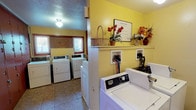 This single-floor shared laundry facility offers a practical and organized space designed to serve multiple users efficiently. The room is finished with warm yellow walls that create a bright and inviting atmosphere, complemented by durable tile flooring throughout.

The laundry area is outfitted with multiple full-sized Maytag top-load washers and dryers, arranged to maximize usability and flow. The machines are divided across two sections of the room, allowing several users to do laundry simultaneously without congestion. Utility hookups are clearly accessible along the walls, and a dedicated shelf above one section provides a convenient surface for personal items, decorated with a basket and floral arrangements that add a welcoming touch.

A standout feature of this facility is the full wall of individual numbered storage lockers finished in a rich red-brown tone. These lockers offer residents dedicated, secure storage space, adding a significant layer of functionality beyond laundry use.

Natural light filters in through a window positioned at the far end of the room, keeping the space feeling open and well-lit alongside the overhead flush-mount lighting. A textured ceiling with a smoke detector and wall-mounted light fixtures rounds out the practical design. Overall, this laundry room is thoughtfully arranged to balance utility, storage, and comfort for everyday use.