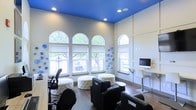 This single-floor space makes an immediate impression with its striking cobalt blue ceiling, which contrasts sharply against warm grey walls and creates a lively, energetic atmosphere. The room is anchored by a row of tall, arched windows that flood the interior with abundant natural light and offer pleasant views of the surrounding greenery outside. The high ceilings amplify the sense of openness and volume throughout the space.

The furnishings strike a balance between functionality and style. Black leather armchairs and tufted white ottomans are arranged to encourage casual gatherings and collaborative interaction, while a sleek, modern standing desk equipped with a desktop computer provides a dedicated workstation along one wall. An ergonomic task chair at an additional desk reinforces the space's dual purpose as both a lounge and a productive work environment.

Decorative blue circular wall accents echo the bold ceiling color and add an artistic dimension to the overall design. The wood-toned flooring grounds the space with warmth and texture, complementing the bright, contemporary palette above. Recessed ceiling lighting supplements the natural light, ensuring the space remains well-illuminated at all times. Overall, this space is thoughtfully designed to be both visually engaging and highly functional.