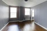 This single-floor space showcases a clean, functional design with a timeless aesthetic. The main room is finished with rich, dark hardwood floors that contrast elegantly against the gray-painted walls, creating a polished yet understated atmosphere. Crown molding runs along the ceiling perimeter, adding a classic architectural detail that elevates the overall character of the space.

Three windows are positioned across two walls, allowing natural light to fill the room from multiple angles. A wall-mounted air conditioning unit is installed beneath the central window, accompanied by a radiator, indicating a dual climate control setup capable of managing both heating and cooling needs year-round. White trim work, including baseboards and window casings, provides crisp contrast against the gray walls.

A ceiling-mounted light fixture provides overhead illumination, complementing the natural light from the windows. A doorway visible along the right wall suggests additional access to an adjoining room or hallway. The space is currently unfurnished, making it easy to assess its versatile layout and the quality of its finishes.