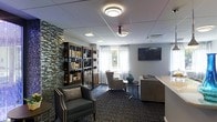 This thoughtfully designed single-floor space functions as a comfortable lounge and library, blending practicality with refined aesthetics. A striking stacked-stone accent wall adds texture and visual depth, serving as an elegant backdrop to a pair of tall, well-stocked dark-framed bookcases that line the corner of the room. The flooring features a richly patterned carpet in deep blue and grey tones, lending warmth and sophistication throughout the space. Seating is arranged to encourage relaxation and conversation, with upholstered armchairs positioned around a sleek metal side table, complemented by a sofa that creates an inviting gathering area. Sheer white curtains frame a window that allows natural light to filter softly into the room. A wall-mounted flat-screen television is positioned prominently, making the space equally suited for entertainment and leisure. Overhead, flush-mount circular light fixtures provide consistent, ambient illumination throughout. The drop ceiling with recessed panels and integrated HVAC contributes to a clean, polished finish. Overall, the space strikes a balance between a classic library atmosphere and a modern communal lounge, offering a welcoming environment for reading, socializing, or unwinding.