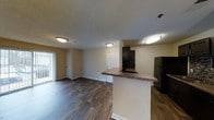 This single-floor space showcases a practical and cohesive open-concept design that seamlessly connects the kitchen and living areas. The kitchen is anchored by a peninsula counter with a dark laminate surface, offering additional workspace and casual seating potential. Dark cabinetry adds a contemporary contrast against the light-toned walls, and black appliances — including a refrigerator and range — complete the kitchen's modern aesthetic. The continuous dark wood-style flooring runs throughout the main living area, lending warmth and visual continuity to the space. A textured ceiling adds subtle character overhead, while flush-mounted light fixtures provide consistent ambient lighting throughout. The living area is generously open and unobstructed, offering flexibility for a variety of furniture arrangements. A sliding glass door along one wall floods the interior with natural light and provides access to an outdoor area, enhancing the sense of openness. White interior doors and clean white baseboards frame the neutral walls, creating a crisp and well-maintained finish. The overall ambiance is bright, functional, and move-in ready, making it a versatile space suited for everyday living.