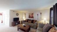 This two-story residence offers a practical and comfortable layout spread across two levels. The main floor opens into a welcoming living room finished with plush carpet, neutral walls, and a textured ceiling. A staircase with white railings connects the levels, and natural light filters in through windows dressed with dark grommet-style curtains. The living area provides ample space for seating and entertainment.

Adjacent to the living room, the kitchen features white cabinetry, granite-look countertops, and white appliances including an electric range with an overhead vent hood. Upper glass-front cabinets add a refined touch. A separate dining area with hardwood-style flooring accommodates a round glass-top table and receives generous natural light through large windows with a decorative valance.

Upstairs, both bedrooms are carpeted and include ceiling fans with light fixtures for year-round comfort. Each room offers window treatments and sufficient space for full bedroom furniture sets. The full bathroom is clean and functional, featuring a single vanity, tub-shower combination, and neutral finishes. A half bath provides added convenience for guests on the main level.