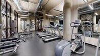 Located at 2925 Dupont Ave S in Minneapolis, MN, this well-equipped fitness center offers a comprehensive exercise environment within a compact yet efficiently utilized space. The room features an industrial-modern aesthetic, highlighted by exposed ductwork running along the ceiling, structural concrete columns, and linear fluorescent lighting that ensures the space is well-lit throughout.

The exercise room is outfitted with a robust selection of cardio equipment, including multiple treadmills, elliptical machines, and a stationary bike, primarily from Precor. A dedicated free weights area provides a full rack of dumbbells alongside kettlebells and medicine balls, catering to strength training needs. Additional equipment includes a rowing machine and a cable/weight machine for versatile workout options.

Large mirrors span the walls, visually expanding the room and allowing users to monitor their form. The flooring consists of durable dark rubber, practical for high-impact activity. Decorative wall panels featuring bold, swirling patterns in brown and gold tones add a distinctive design element, complementing the warm neutral wall tones. The entrance is framed by an elegant wood door with matching decorative glass panels. Overall, the space is thoughtfully designed to balance functionality with a polished, modern aesthetic.