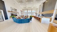 The Alexander Clubhouse is a thoughtfully designed single-floor community space that seamlessly blends comfort, functionality, and modern style across a generous footprint. The heart of the facility is a dramatic open-plan living and kitchen area defined by soaring coffered ceilings, large windows that flood the space with natural light, and wide-plank light wood flooring that flows throughout. The living area is elegantly furnished with bold blue sectional seating, curated artwork, and a fireplace with a wood-framed surround, creating an inviting atmosphere for gathering. A stunning kitchen bar anchors one side of the space, featuring a long white marble countertop, warm wood cabinetry with fluted detailing, a textured dark tile backsplash, and built-in double ovens. An adjacent living room offers additional seating with high ceilings and glass entry doors. A separate dining area showcases a striking live-edge wood resin table paired with contemporary abstract artwork and direct access to a private office equipped with a built-in marble-topped desk, wood storage drawers, and wall-mounted lighting. The well-equipped fitness center offers a wide selection of cardio and strength training machines, rubber flooring, recessed lighting, and outdoor views through a row of windows.