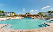 Located at 16100 Myriad Ln in Fort Myers, FL, this expansive outdoor amenity area offers a resort-style experience within a residential community. At its centerpiece is a generously sized swimming pool with clear turquoise water, a classic geometric shape, and entry steps for easy access. The pool deck is finished in warm stone-toned pavers and is lined with lounge seating, providing ample space for sunbathing and relaxation beneath the Florida sun. A decorative gazebo anchors one end of the pool area, while lush tropical landscaping — including tall palm trees and manicured plantings — frames the surroundings throughout.

The covered outdoor lounge area is a standout feature, boasting soaring vaulted ceilings with ceiling fans, large-format tile flooring, and floor-to-ceiling sliding glass doors that open to adjacent interior spaces. This shaded zone is furnished with wicker sectional seating in vibrant turquoise upholstery, a dining table and chairs, and wall-mounted televisions for outdoor entertainment. A separate covered corridor provides additional recreational space, including a ping pong table, decorative ceramic urns, and open-air views of the pool. Scenic water views and community buildings are visible in the background, completing the cohesive, tropical ambiance of this well-appointed outdoor retreat.