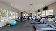This fitness center is a comprehensively outfitted exercise facility designed to support a wide range of workout routines. The space is organized into distinct functional zones, beginning with a dedicated cardio area that houses multiple True-brand treadmills, elliptical trainers, and a stationary bike, each positioned in front of large windows that frame views of the surrounding greenery and outdoor environment. Wall-mounted flat-screen televisions are positioned throughout for entertainment during workouts.

The strength training area features a multi-station cable machine along with adjustable benches and stability balls, providing versatile options for resistance and functional fitness exercises. A full-length mirrored wall spans one side of the space, visually expanding the room and allowing users to monitor their form. The mirrors also reflect natural light streaming in from the numerous windows, keeping the interior bright and energizing throughout the day.

Durable rubber flooring covers the entire facility, offering both safety and comfort underfoot. Recessed lighting supplements the natural light, ensuring consistent illumination at all hours. A set of glass-paned double doors provides a welcoming entrance, while a pass-through window connects to an adjacent lounge area. The neutral walls and clean architectural lines give the space a polished, professional ambiance suited for daily fitness use.