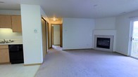 This single-floor residence offers a straightforward and functional layout centered around a spacious living area. The room is finished with wall-to-wall carpeting in a neutral gray-beige tone, contributing to a quiet, comfortable atmosphere throughout. White walls keep the space feeling open and clean, providing a versatile backdrop for a variety of furnishing styles.

A built-in fireplace with a black glass front is situated along one wall of the living area, serving as a practical focal point. The fireplace surround is finished in a simple white material that blends seamlessly with the surrounding walls.

A hallway visible from the main living space leads to additional rooms, with wood-trimmed doorframes providing a warm contrast to the white walls. Closet doors line the hallway, suggesting generous storage capacity. Flush-mount ceiling lighting in the hallway provides adequate illumination to the interior corridor.

The overall design is clean and understated, with consistent finishes and neutral tones throughout. The layout appears efficient, with the hallway connecting the living area to the private spaces of the home. The space is well-suited for those seeking a low-maintenance, move-in-ready environment.