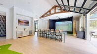 Located at 4711 Azalea Landing Dr in Wilmington, NC, this single-floor amenity facility offers a thoughtfully designed and well-appointed communal space. The layout encompasses two dining areas, an exercise room, and a dedicated golf simulator bay, all unified by light-toned hardwood-style flooring and crisp white walls throughout.

The golf simulator lounge is a standout feature, anchored by a full-screen simulator bay with artificial turf and draped black curtain enclosure. Exposed wooden beam trusses soar overhead, lending warmth and architectural character to the vaulted ceiling. A bar-height counter with cane-back seating faces the simulator, while a built-in wet bar and beverage refrigerator serve the dining areas nearby. A glass garage door opens to an outdoor pool area, blurring the line between indoor and outdoor living.

The exercise room is spacious and bright, featuring large glass garage doors that invite natural light and outdoor views. Ceiling fans keep the air circulating above a fleet of cycling bikes and strength equipment. A striking living moss accent wall, adorned with a neon motivational sign and mounted display screens, creates an energetic focal point.

A boldly wallpapered entry vestibule with herringbone tile flooring and black-framed glass doors provides a stylish welcome. Throughout, the design balances modern sophistication with inviting, resort-inspired touches.