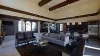 This single-story clubhouse is designed to impress from the moment you step inside. The main lounge area commands attention with its soaring ceilings adorned with rich, dark-stained exposed wood beams that lend a warm, architectural character to the space. Large arched windows along one wall flood the interior with abundant natural light, creating a bright and welcoming atmosphere. The lounge is furnished with a generous gray tufted sofa and leather armchairs, all arranged around a sleek round metallic coffee table atop a richly patterned area rug — offering a sophisticated yet comfortable gathering space. Decorative circular wall accents and framed artwork add thoughtful finishing touches throughout. Adjacent to the lounge, a business or activity area features workstations with computer monitors, providing a functional resource for residents. A stylishly appointed bar or counter area with dark tile backsplash and pendant lighting serves as an additional focal point, perfect for social gatherings. The open-concept layout ensures seamless flow between each zone, while the cohesive neutral and dark toned color palette ties the entire space together into a polished, upscale clubhouse environment.