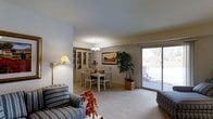 This single-floor residence offers a warm and inviting layout centered around a seamlessly connected living and dining area. The living space is furnished with comfortable striped upholstered seating and accented by a wall-mounted swing-arm lamp, creating a cozy and functional atmosphere. A vibrant floral arrangement adds a pop of color to the space.

The dining area is positioned adjacent to the living room and features a classic white dining set with ladder-back chairs, centered beneath an elegant chandelier. A traditional china cabinet with glass-front display shelving adds a refined, timeless touch to the room, while a framed landscape painting on the wall contributes to the warm, homey aesthetic.

A large sliding glass door floods the space with natural light and offers a direct view to the exterior, where mature trees and a glimpse of the surrounding neighborhood can be seen. A tall potted plant near the door brings a touch of greenery indoors, softening the transition between interior and exterior.

The interior is finished with neutral wall tones and plush carpeting throughout, lending the space a cohesive and comfortable feel. The overall ambiance is classic and functional, with thoughtful details that make this single-level home both practical and pleasant to inhabit.