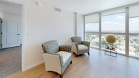 This single-floor residence offers a thoughtfully designed layout that balances comfort and functionality. The living area is a standout feature, showcasing expansive floor-to-ceiling windows that flood the space with natural light and frame sweeping panoramic views of the surrounding cityscape. Light wood-tone flooring runs throughout the main living areas, enhancing the open, airy feel of the space. The seating area accommodates multiple lounge chairs and a sofa, all oriented to take full advantage of the impressive views.

The kitchen is efficiently arranged in an L-shaped configuration and is outfitted with warm wood cabinetry, granite countertops, and stainless steel appliances including a refrigerator, microwave, and dishwasher. Recessed lighting and tile flooring complete the functional yet attractive cooking space.

The bedroom is generously proportioned, featuring plush carpeting, clean white walls, and large windows dressed with floor-length drapes that allow for both light and privacy. The room offers ample space for furnishings and connects seamlessly to the rest of the unit.

The bathroom is well-sized and finished with warm wood vanity cabinetry, a light stone-toned tile floor, a glass-enclosed shower, and clean white fixtures. Together, these spaces create a cohesive, modern living environment with striking elevated views as a defining characteristic.
