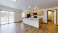 This single-floor unit offers a clean, functional layout with consistent warm-toned wood vinyl flooring and neutral cream walls throughout. The heart of the home is an open-concept kitchen and living area, where a central island with a light countertop and built-in sink serves as the focal point. Light maple cabinetry lines the kitchen walls, providing generous storage alongside tall pantry-style cabinets. A full suite of black appliances — including a refrigerator, electric range, and over-the-range microwave — lends a cohesive, modern look. The kitchen flows seamlessly into the living space, which benefits from abundant natural light via a large sliding glass door that opens to an outdoor balcony and an additional window overlooking the exterior. The bedroom features a ceiling fan with light, a wall-mounted HVAC unit, and a window offering views of surrounding trees, creating a calm and comfortable retreat. Additional rooms display the same durable flooring and clean, minimalist finishes. The bathroom is equipped with a walk-in shower unit, brushed nickel towel bar, toilet paper holder, and a wood-toned vanity, all finished with the same continuity seen throughout the space.