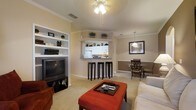 This single-story residence offers two bedrooms and two bathrooms within a thoughtfully designed floor plan. The living area serves as the heart of the home, featuring warm neutral-toned walls, crown molding, and plush carpeting underfoot. A ceiling fan provides both comfort and ambient lighting, while a built-in shelving unit along one wall offers display and storage space, complementing the entertainment setup.

The open-concept design connects the living area seamlessly to a breakfast bar, which separates the kitchen from the main living space. The kitchen features white cabinetry, standard appliances, and an overhead shelf adorned with greenery, adding a touch of warmth. Bar-height seating at the counter creates a casual dining option.

Adjacent to the living area, a dedicated dining space accommodates a small table and chairs, defined by wainscoting detail along the lower portion of the wall, adding a refined architectural touch. The overall ambiance is warm and inviting, with consistent neutral tones, coordinated finishes, and a layout that balances open communal areas with defined functional zones throughout the home.