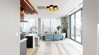 This contemporary amenity lounge spans a generous single floor, offering a thoughtfully designed open-plan layout that seamlessly blends social, dining, and lounge functions. Floor-to-ceiling glass walls and expansive windows flood the space with natural light while framing sweeping panoramic city skyline views, creating a vibrant, elevated atmosphere.

The kitchen area serves as a central anchor, featuring a large curved white quartz island with seating for multiple guests, professional-grade appliances, and a dramatic wood-slat ceiling detail with integrated lighting overhead. A dedicated wall of shelving in warm wood tones adds a curated, library-like character to one corner, complemented by a media wall and colorful accent furnishings.

The lounge area is appointed with plush, modern seating arranged around a patterned area rug, while sliding glass doors open directly onto an outdoor terrace, extending the usable space and maximizing the connection to the urban landscape beyond. A long communal dining table positioned near the perimeter windows provides an ideal setting for collaborative gatherings or casual dining.

Light-toned wood flooring runs throughout, unifying the various zones, while exposed concrete columns and the striking slatted wood ceiling installation add architectural texture and warmth to the overall design.