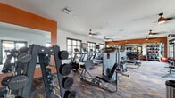 This fitness center is designed to deliver a complete workout experience within a single, open-plan floor. The space is defined by a bold color palette, pairing warm terracotta-orange accent walls with crisp white ceilings and dark-framed windows that invite generous natural light and outdoor views. Textured, multi-tone carpet tiles in charcoal, gold, and grey add visual depth underfoot while providing a durable, functional surface throughout.

The cardio zone is well-stocked with a row of treadmills, ellipticals, and a stair climber, all positioned along the windowed walls to maximize natural light during workouts. Ceiling fans overhead contribute to comfortable air circulation. A dedicated strength training area features a full dumbbell rack spanning a wide mirrored wall, which enhances the sense of space, alongside multiple selectorized cable and weight machines from brands including TKO and True.

Near the entrance, a tiled accent wall houses a dual water fountain and bottle-filling station, complemented by a pendant light and a glass door that connects to the exterior. The layout flows efficiently between zones, making the facility feel spacious, organized, and well-suited for a diverse range of fitness routines.