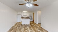 This efficiently designed single-floor unit offers a thoughtful layout across a compact footprint, accommodating a combined kitchen and living area, one bedroom, a full bathroom, and two hallways. Wood-look plank flooring runs consistently throughout the entire unit, providing a cohesive and warm aesthetic in every room.

The open-plan kitchen and living area serves as the central hub of the unit. The kitchen features white cabinetry extending to the upper walls, granite-style laminate countertops, and a peninsula with additional counter space and open shelving below—ideal for dining or food preparation. White appliances, including a refrigerator, electric range, microwave, and dishwasher, are neatly integrated. A ceiling fan with a light fixture serves both the kitchen and living area, which receives natural light through windows fitted with blinds and is anchored by a distinctive red entry door.

The bedroom is a well-proportioned, sunlit room with a ceiling fan, a window with outdoor views, and clean white walls. The bathroom is finished with white cabinetry, a vanity with a mirror, a combination shower-tub unit, and consistent wood-look flooring, maintaining the unit's uniform design language throughout.