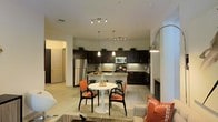 This single-floor residence features an open-concept layout that seamlessly connects the kitchen, dining, and living areas into one cohesive living environment. The kitchen is appointed with dark espresso cabinetry that extends to the upper walls, providing generous storage. A tiled backsplash runs beneath the upper cabinets, complementing the granite-style countertops. Stainless steel appliances, including a full-size refrigerator and range, are integrated throughout. A peninsula with pendant lighting serves as both a prep space and casual bar seating with black stools. Track lighting overhead ensures ample illumination across the kitchen. The dining area is anchored by a round white pedestal table surrounded by black-framed chairs with warm orange cushions, adding a vibrant accent against the neutral palette. Light wood-toned flooring flows throughout, contributing to the open, airy feel of the space. The living area, visible in the foreground, continues the warm color scheme with coordinating soft furnishings. A tall arched floor lamp adds a design focal point near the transition between spaces. A hallway leading to additional rooms is visible toward the rear left of the space.