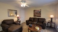 This single-floor space offers a comfortable and cohesive living environment designed with both function and style in mind. The living room serves as the heart of the space, featuring plush dark-upholstered seating including a sofa and matching armchair arranged around a warm, round wooden coffee table. A ceiling fan with integrated lighting provides both comfort and illumination, keeping the room well-ventilated and bright. The neutral-toned walls provide a versatile backdrop, accented by decorative artwork that adds a lively, urban character to the room. A classic table lamp and floral arrangement on a side table contribute to the warm, welcoming ambiance. Carpet flooring adds a layer of softness underfoot, enhancing the cozy atmosphere of the living area. A closet or interior door is accessible from the main living space, suggesting thoughtful storage and room connectivity. The overall design aesthetic is traditional and approachable, with rich wood tones and dark upholstery creating a grounded, harmonious interior. The space is well-suited for relaxed everyday living, offering a layout that balances comfort with practicality in a compact, efficiently organized single-level floor plan.