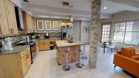 This lounge room is designed as a welcoming communal space that seamlessly integrates a fully equipped kitchen with an open, comfortable gathering area. The kitchen is outfitted with warm, natural wood cabinetry, stainless steel appliances including a gas range and refrigerator, and a decorative tile backsplash. A large central island with a neutral countertop provides additional workspace and is complemented by modern chrome bar stools, making it an ideal spot for casual dining or socializing. Pendant lighting above the island adds a focused, stylish touch.

The living area is anchored by a striking floor-to-ceiling stacked stone fireplace, which also houses a wall-mounted television, serving as the room's focal point. Mid-century modern-style armchairs and sofas in bold amber tones are arranged around the fireplace, creating an inviting seating arrangement. Stacked stone columns echo the fireplace's design, reinforcing a cohesive aesthetic throughout the space.

Large windows along the perimeter flood the interior with natural light and offer views of an outdoor patio area. Light-toned tile flooring with a subtle herringbone accent rug grounds the lounge area. Colorful framed artwork and fresh floral arrangements add warmth and personality to the overall space.