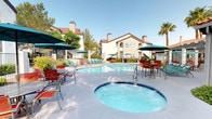 This outdoor amenity space showcases a beautifully maintained resort-style pool area designed for relaxation and leisure. The centerpiece is a generously sized, freeform swimming pool with crystal-clear turquoise water, complemented by a separate circular spa ideal for unwinding. The expansive surrounding sundeck is finished in smooth concrete and outfitted with teal lounge chairs, vibrant red seating sets, and teal market umbrellas, providing ample space for sunbathing and socializing. A covered pergola structure adjacent to the pool area offers a shaded retreat, perfect for gatherings out of direct sun. The space is framed by mature palm trees, deciduous trees, and well-kept shrubs and hedges that add lush greenery and a sense of privacy. The backdrop features multi-story residential buildings with a distinct Mediterranean architectural style, characterized by stucco exteriors, terra-cotta tile rooflines, arched windows, and decorative chimneys. Wrought iron fencing and gated access points contribute to the refined aesthetic. The overall ambiance is bright, open, and inviting, evoking the feel of a private resort with well-considered details throughout the outdoor environment.
