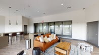 This single-floor amenity space is designed with a contemporary aesthetic, blending industrial and modern elements throughout its open layout. Wood-look tile flooring runs continuously across the space, anchoring each distinct zone while maintaining a cohesive visual flow. Recessed lighting and stylish pendant fixtures illuminate the interior, complementing the neutral white walls and dark-framed architectural accents.

The lounge area features a rich cognac leather sofa layered with accent pillows, positioned around a wood-and-metal coffee table atop a colorful area rug. Adjacent to the lounge is a bar-height counter island with dark countertops and leather bar seating, suspended pendant lights above, and a beverage refrigerator nearby — ideal for social gatherings.

A standout feature is the dramatic exposed brick accent wall, which rises to soaring ceilings and frames an upright piano set on a raised platform. A wall-mounted television and curated Nashville artwork add character to this area. Large sliding glass doors open directly onto an outdoor balcony, flooding the space with natural light and offering expansive views of the surrounding treetops and skyline.

The game zone includes a full-size pool table beneath a pendant light, while a shuffleboard table near the entrance adds further recreational appeal. The overall ambiance is vibrant yet refined.