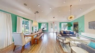 Located at 5555 North Military Trail in Boca Raton, Florida, this single-floor space offers an open, airy layout thoughtfully designed for both socializing and relaxation. The interior is immediately distinguished by its bold teal accent walls, which contrast elegantly against crisp white ceilings and trim, creating a fresh, energetic atmosphere throughout.

The space is divided into two seamlessly connected functional areas. The dining area features a substantial wooden farm table paired with bench seating and upholstered chairs, suspended beneath a large cylindrical pendant light. Multiple double-hung windows dressed in flowing white curtains line the walls, bathing the area in generous natural light and offering views of the lush greenery outside.

The living area is anchored by a striking gold sputnik chandelier that draws the eye upward to the vaulted ceiling, adding a touch of mid-century modern glamour. Comfortable gray sofas and accent chairs are arranged around a glass-topped coffee table, complemented by a wall-mounted television and curated décor. A set of French doors provides direct access to the outdoors, further enhancing the bright, open feel.

Rich hardwood flooring flows continuously throughout, tying both areas together cohesively. A dedicated workspace with a desk and chair is also integrated into the layout, adding functional versatility to this well-appointed, light-filled space.