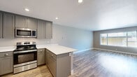 Located at 28500 Mission Blvd in Hayward, California, this well-appointed one-bedroom, one-bathroom unit offers a thoughtfully designed single-level layout with modern finishes throughout. The open-concept kitchen and living area serves as the heart of the home, featuring grey shaker-style cabinetry, crisp white countertops, a peninsula with bar seating, and a full suite of stainless steel appliances including a refrigerator, dishwasher, microwave, and range. Recessed lighting and wood-style plank flooring tie the kitchen and living spaces together seamlessly. The living room is generously sized, bathed in natural light from large windows, and provides direct access to a private balcony through a distinctive teal-framed glass door. The bedroom is carpeted for comfort and features a wide window overlooking a green outdoor setting, creating a calm and restful atmosphere. A dedicated closet provides practical storage. The bathroom is well-equipped with a full tub/shower combo, grey vanity with white countertop, a large mirror, and matching wood-style flooring. An in-unit washer is also present, adding everyday convenience to this clean, move-in-ready home.