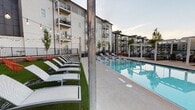 Located at Cosby Village in Chesterfield, Virginia, this thoughtfully designed outdoor amenity area serves as the social centerpiece of the community. The standout feature is a generously sized resort-style swimming pool with crystal-clear blue water, flanked by a wide pool deck equipped with modern lounge chairs and sleek white pergola structures that provide shaded seating areas. Neatly planted trees and manicured landscaping add a refined, park-like quality to the surroundings.

Adjacent to the pool, a dedicated sunbathing lawn features rows of contemporary chaise lounges arranged on lush artificial turf, accented by warm globe string lights strung overhead — creating an inviting ambiance for both daytime relaxation and evening gatherings. Vibrant orange accent chairs punctuate the greenery, adding a pop of color to the carefully curated outdoor palette.

A separate courtyard entrance area showcases a striking brick and dark-panel building exterior, adorned with a bold decorative mural and additional overhead string lighting, lending an urban, neighborhood feel to the community. Wrought-iron fencing, brick accent walls, and well-maintained plantings frame the walkways throughout. The multi-story residential buildings surrounding the amenity space feature private balconies that overlook the pool and grounds, reinforcing the community's cohesive and upscale character.