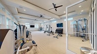This generously sized fitness center offers a comprehensive workout facility spread across a single open floor. The space is designed with a clean, modern aesthetic, featuring tray ceilings accented with decorative molding and multiple ceiling fans that promote air circulation throughout the room. Light-toned flooring spans the main workout area, contributing to the bright and airy atmosphere created by an abundance of natural light streaming through large windows and clerestory windows positioned along the upper walls.

The facility is thoughtfully divided into distinct training zones. A dedicated cardio section is equipped with multiple treadmills, a stair climber, rowing machine, and stationary bikes, all positioned adjacent to wide windows that provide pleasant outdoor views during workouts. The strength training area features a comprehensive selection of cable machines, multi-functional cable systems, and weight benches, alongside a well-stocked dumbbell rack. A separate enclosed room with floor-to-ceiling glass partition walls houses additional strength and resistance equipment, offering a more private workout environment.

Multiple wall-mounted flat-screen televisions are positioned throughout the space for entertainment. The combination of ample natural light, thoughtful equipment layout, and modern architectural details makes this fitness center a highly functional and inviting facility.
