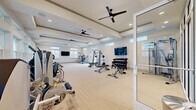 This generously sized fitness center offers a comprehensive workout facility spread across a single open floor. The space is designed with a clean, modern aesthetic, featuring tray ceilings accented with decorative molding and multiple ceiling fans that promote air circulation throughout the room. Light-toned flooring spans the main workout area, contributing to the bright and airy atmosphere created by an abundance of natural light streaming through large windows and clerestory windows positioned along the upper walls.

The facility is thoughtfully divided into distinct training zones. A dedicated cardio section is equipped with multiple treadmills, a stair climber, rowing machine, and stationary bikes, all positioned adjacent to wide windows that provide pleasant outdoor views during workouts. The strength training area features a comprehensive selection of cable machines, multi-functional cable systems, and weight benches, alongside a well-stocked dumbbell rack. A separate enclosed room with floor-to-ceiling glass partition walls houses additional strength and resistance equipment, offering a more private workout environment.

Multiple wall-mounted flat-screen televisions are positioned throughout the space for entertainment. The combination of ample natural light, thoughtful equipment layout, and modern architectural details makes this fitness center a highly functional and inviting facility.