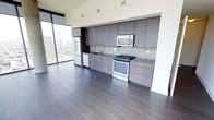 This contemporary high-rise unit showcases a refined, open-concept layout with a seamless flow between the kitchen and living areas. The kitchen is the centerpiece of the space, featuring floor-to-ceiling flat-panel cabinetry in a warm grey-brown finish, complemented by a neutral tile backsplash and dark stone countertops. Stainless steel appliances — including a refrigerator, electric range, built-in microwave, and dishwasher — provide a sleek, modern aesthetic. Rich, dark hardwood flooring runs throughout, grounding the space with warmth and contrast against the light-toned walls and cabinetry. Large floor-to-ceiling windows flood the interior with abundant natural light and frame impressive panoramic views of the surrounding city skyline, enhancing the sense of openness. The clean lines, minimalist design, and high-quality finishes throughout reflect a thoughtfully curated modern aesthetic. The expansive glazing not only maximizes light but also creates a strong visual connection to the vibrant urban landscape beyond, making this a compelling space that balances functional design with elevated city living.