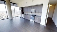 This contemporary high-rise unit showcases a refined, open-concept layout with a seamless flow between the kitchen and living areas. The kitchen is the centerpiece of the space, featuring floor-to-ceiling flat-panel cabinetry in a warm grey-brown finish, complemented by a neutral tile backsplash and dark stone countertops. Stainless steel appliances — including a refrigerator, electric range, built-in microwave, and dishwasher — provide a sleek, modern aesthetic. Rich, dark hardwood flooring runs throughout, grounding the space with warmth and contrast against the light-toned walls and cabinetry. Large floor-to-ceiling windows flood the interior with abundant natural light and frame impressive panoramic views of the surrounding city skyline, enhancing the sense of openness. The clean lines, minimalist design, and high-quality finishes throughout reflect a thoughtfully curated modern aesthetic. The expansive glazing not only maximizes light but also creates a strong visual connection to the vibrant urban landscape beyond, making this a compelling space that balances functional design with elevated city living.