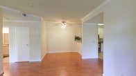 This single-floor residence offers a clean, open layout designed for comfortable everyday living. The main living area is anchored by warm hardwood flooring that runs throughout the space, complemented by crisp white walls and elegant crown molding that add a polished, classic character to the interior. A ceiling fan with a light fixture provides both comfort and ambient illumination, making the room feel airy and well-lit.

The living area flows naturally into adjacent spaces through open archways and wide doorways, creating a sense of connected, open-plan living. A decorative ledge provides a subtle architectural detail, offering a spot for greenery or accents. To one side, a passthrough or transitional area leads toward additional rooms, while an opening on the opposite side reveals the kitchen, which features white cabinetry, dark countertops, and stainless steel appliances. The kitchen is bright and functional, with ample cabinet storage.

The consistent use of white tones across walls, trim, and cabinetry creates a cohesive, light-filled atmosphere throughout the home. The crown molding and clean architectural lines lend the space a timeless, well-finished quality that makes it both inviting and functional.