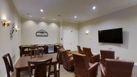 This single-floor entertainment lounge is thoughtfully designed as a private screening and social gathering space. The room is anchored by a large wall-mounted flat-screen television, making it ideal for movie viewings and group entertainment. An array of plush, high-back brown leather armchairs are arranged to face the screen, offering comfortable seating for an audience, while additional high-top tables and chairs provide a more casual dining or social option along the perimeter.

A playful "It's Showtime" marquee-style wall sign adds a cinematic character to the space, reinforcing its theater-inspired ambiance. Along one wall, a well-appointed wet bar area features a granite countertop, cabinetry, a sink, and a compact under-counter refrigerator — perfect for serving refreshments during screenings or events.

The room is finished with warm neutral walls, crown molding, and recessed ceiling lighting that creates a soft, inviting atmosphere. Wall sconces add an elegant accent and contribute to the layered lighting design. The light-toned carpet and classic trim work lend a refined, comfortable feel throughout. A solid paneled door with a sleek smart-panel entry system ensures privacy and security for this well-equipped amenity space.