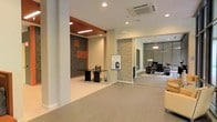 This single-floor commercial space presents a well-designed, open layout that seamlessly integrates multiple functional zones within a contemporary framework. The interior is finished with light-toned flooring that runs throughout, creating a cohesive and clean aesthetic across the entire space.

A dedicated workstation area features a sleek dark desk set against a bold accent wall clad in textured stone tiles with orange geometric detailing, adding a striking visual focal point. Recessed ceiling lighting and a cassette-style HVAC unit overhead ensure comfort and even illumination throughout.

A natural stacked-stone column serves as an architectural divider, guiding the eye toward a secondary lounge or meeting area. This adjoining zone is enclosed with floor-to-ceiling glass partitions, allowing visibility while maintaining separation. It is furnished with dark seating, a side table, and decorative accents, creating a refined and professional atmosphere.

The open central floor area provides flexible space, currently accommodating a presentation easel, suggesting the layout supports a variety of uses. Overall, the space combines modern materials, thoughtful zoning, and a neutral palette to create a functional and polished commercial environment.