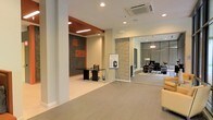 This single-floor commercial space presents a well-designed, open layout that seamlessly integrates multiple functional zones within a contemporary framework. The interior is finished with light-toned flooring that runs throughout, creating a cohesive and clean aesthetic across the entire space.

A dedicated workstation area features a sleek dark desk set against a bold accent wall clad in textured stone tiles with orange geometric detailing, adding a striking visual focal point. Recessed ceiling lighting and a cassette-style HVAC unit overhead ensure comfort and even illumination throughout.

A natural stacked-stone column serves as an architectural divider, guiding the eye toward a secondary lounge or meeting area. This adjoining zone is enclosed with floor-to-ceiling glass partitions, allowing visibility while maintaining separation. It is furnished with dark seating, a side table, and decorative accents, creating a refined and professional atmosphere.

The open central floor area provides flexible space, currently accommodating a presentation easel, suggesting the layout supports a variety of uses. Overall, the space combines modern materials, thoughtful zoning, and a neutral palette to create a functional and polished commercial environment.