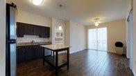 This single-floor residence features an open-concept layout that seamlessly connects the kitchen and living areas, creating a cohesive and functional living space. The kitchen is appointed with rich, dark espresso cabinetry that provides a striking contrast against the light granite countertops. A freestanding kitchen island with a matching granite surface and lower storage shelf serves as both a practical workspace and a natural dividing point between the kitchen and the main living area. A pendant light fixture hangs above the island, adding a focused decorative accent. The kitchen also includes a stainless steel sink and built-in appliances. Rich, dark wood-look flooring runs throughout the main living area, lending warmth and visual depth to the space. The living area is generously sized and bathed in natural light streaming through large floor-to-ceiling sliding glass doors fitted with vertical blinds, offering access to an outdoor area and views of the surrounding greenery. A ceiling fan with integrated lighting is positioned above the living space for added comfort. A glimpse through an interior doorway reveals a bedroom with upholstered headboard detailing, completing this well-designed and light-filled residence.