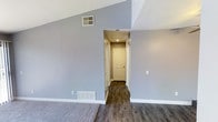 This upgraded 1-bedroom, 1-bathroom unit offers a well-designed single-floor layout that balances functionality with modern style. The living area immediately impresses with its vaulted ceilings, which lend an open, airy feel to the space and create a sense of expansiveness beyond its footprint. Soft, neutral gray walls complement the crisp white trim and baseboards throughout, providing a clean and contemporary aesthetic. Dark hardwood-style flooring runs through the main living and hallway areas, adding warmth and a polished look, while plush carpeting in the living space offers added comfort underfoot. A well-lit hallway leads to the entryway, where a classic six-panel door with recessed overhead lighting provides a welcoming transition into the home. The layout includes practical floor-level vents and wall-mounted climate controls, ensuring year-round comfort. The overall ambiance is bright, neutral, and move-in ready, making it an ideal space for those seeking a modern, upgraded living environment with thoughtful architectural details and quality finishes throughout.