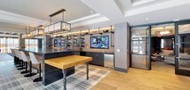 This thoughtfully designed single-floor amenity space delivers a sophisticated, multi-functional environment that seamlessly blends social and relaxation areas. Wide-plank hardwood flooring runs throughout, tying together each distinct zone with warmth and continuity.

The lounge area offers an inviting arrangement of contemporary chairs in varied styles — from sleek molded seats to plaid-upholstered accent chairs — set against deep navy walls. Large windows with slatted blinds fill the space with natural light and provide views of the surrounding exterior, while coffered ceiling details add architectural depth overhead.

Adjacent to the lounge, a well-appointed bar area serves as a central social hub. It features industrial-style open shelving, a large flat-screen TV mounted against an exposed brick accent wall, a butcher-block table, and a stainless steel under-counter refrigerator. A chain-hung pendant light fixture anchors the space, complemented by ambient LED lighting along the shelving.

A dedicated dining area rounds out the layout, offering rich caramel-toned leather booth seating arranged along a dark accent wall adorned with a curated gallery of framed black-and-white artwork. The overall ambiance is modern and refined, balancing bold design choices with comfortable, functional furnishings suited for gathering, dining, or unwinding.