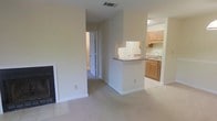 This single-story space is designed with an open, functional layout that connects the main living area to the kitchen through a peninsula-style counter. The living area is carpeted and anchored by a fireplace set into the wall, adding a practical focal point to the room. Neutral, light-colored walls throughout contribute to a clean, airy feel.

The kitchen is positioned adjacent to the living space and is partially separated by a raised countertop peninsula, which provides additional workspace and a natural division between the two areas. The kitchen features medium-toned wood cabinetry, a sink beneath a window, and tile flooring that contrasts with the carpeted living space. Upper and lower cabinets offer ample storage. A range hood is mounted beneath the upper cabinetry.

A hallway visible from the living area leads to additional rooms, suggesting the layout extends further into the unit. Ceiling-mounted HVAC vents are present, indicating a centralized climate control system. The overall ambiance is understated and neutral, offering a versatile, move-in-ready environment well-suited for a variety of furnishing styles.