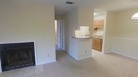 This single-story space is designed with an open, functional layout that connects the main living area to the kitchen through a peninsula-style counter. The living area is carpeted and anchored by a fireplace set into the wall, adding a practical focal point to the room. Neutral, light-colored walls throughout contribute to a clean, airy feel.

The kitchen is positioned adjacent to the living space and is partially separated by a raised countertop peninsula, which provides additional workspace and a natural division between the two areas. The kitchen features medium-toned wood cabinetry, a sink beneath a window, and tile flooring that contrasts with the carpeted living space. Upper and lower cabinets offer ample storage. A range hood is mounted beneath the upper cabinetry.

A hallway visible from the living area leads to additional rooms, suggesting the layout extends further into the unit. Ceiling-mounted HVAC vents are present, indicating a centralized climate control system. The overall ambiance is understated and neutral, offering a versatile, move-in-ready environment well-suited for a variety of furnishing styles.